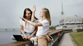 Two Tech women sitting on a concrete ledge by the water,  using a laptop and the phone in USA.
