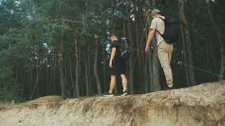 Two white hikers walking along forest edge beside eroded sand ledge, measured pace and backpacks