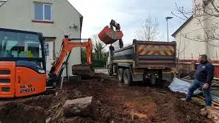 Digger moves spoil heap for grab lorry to pick up while Romesh lookalike watches.