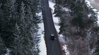 Aerial top down view of a black SUV driving on a remote asphalt road through a snowy evergreen