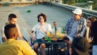 Cheerful friends are chatting and drinking lemonade sitting on rooftop on warm sunny day with snacks