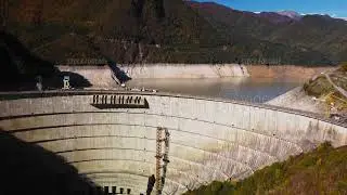 High concrete arch dam holding river located among green mountains filmed by drone